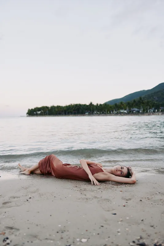 Relaxed woman lying on the sandy beach near calm sea water with distant green hills and soft pastel sky at sunset.