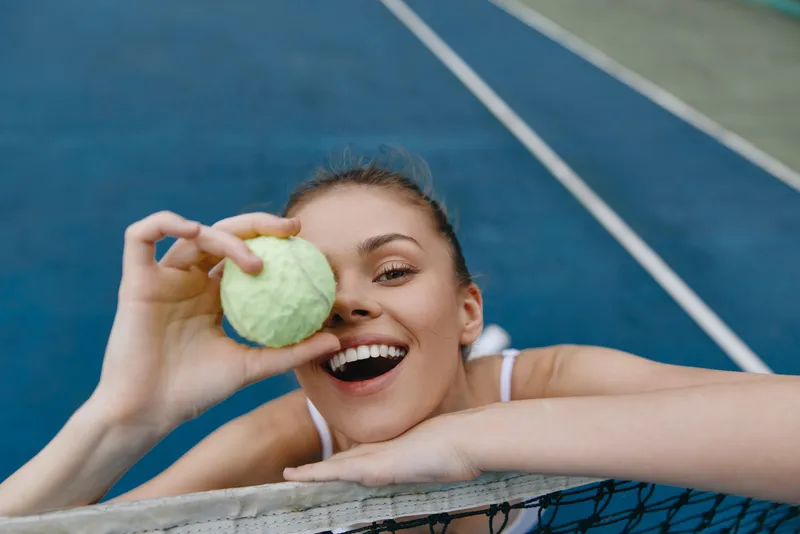 Young woman playing tennis, holding a bright green tennis ball with a joyful smile, sporting athletic wear on a blue tennis court background