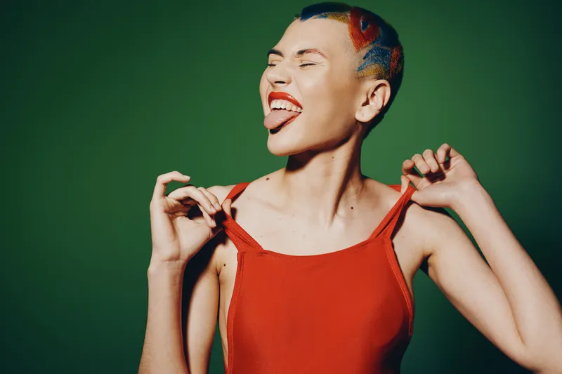 playful woman with colorful short hair in red top showing tongue and pulling straps against green background, expressing fun and bold attitude, vibrant and youthful portrait