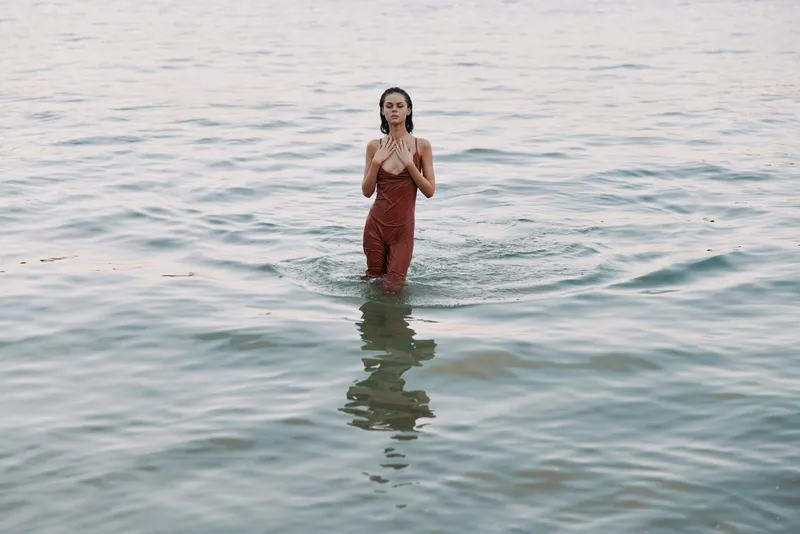 Woman standing in calm sea water wearing a red swimsuit, facing forward, with gentle ripples around her and a reflective surface creating a serene seaside moment.