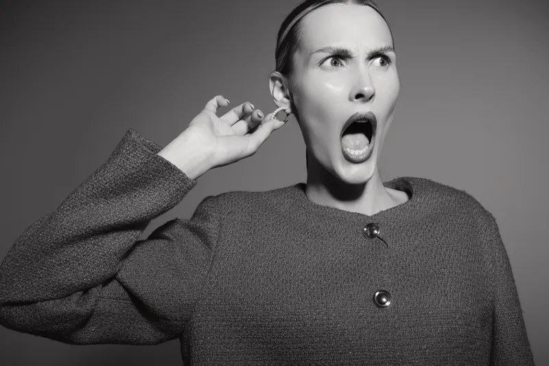 Surprised Caucasian woman with fair skin reacts with open mouth and raised hand near ear. She wears a textured jacket in a studio with dramatic lighting. Concept of shock and unexpected news.