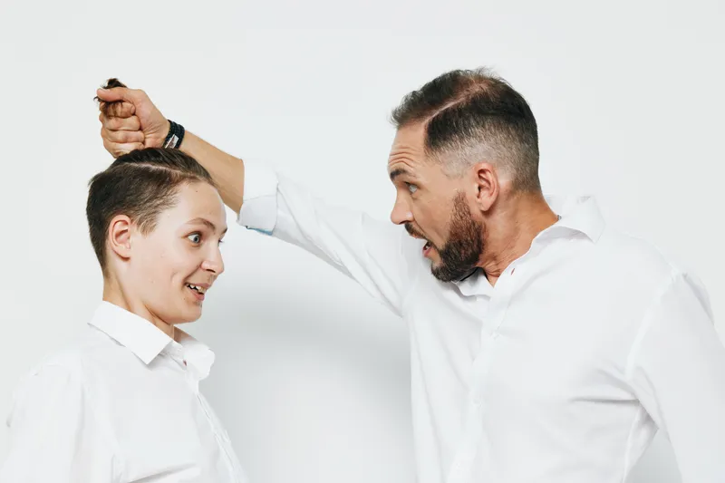 A professional business man interacts with a younger colleague showing playful intensity; he smiles widely while lifting hair in a light office pose against an isolated white background, conveying