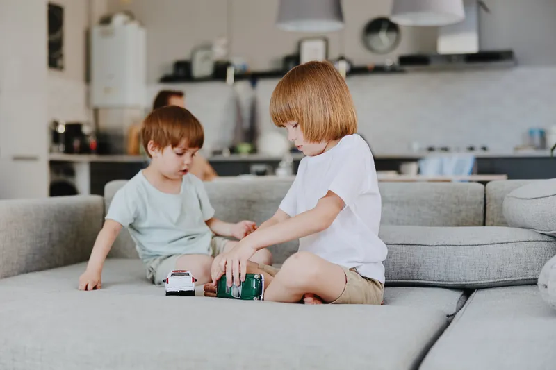 Two boys playing with toy cars on a cozy sofa in a modern living room, showcasing creativity and fun in a bright and welcoming environment.