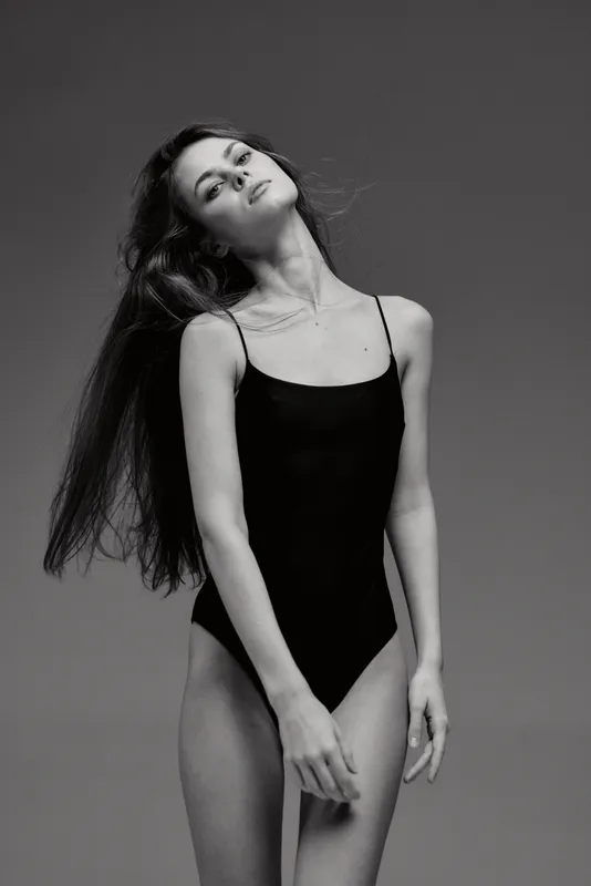 Black and white portrait of a slender woman in a simple black bodysuit posing gracefully with long hair and soft expression