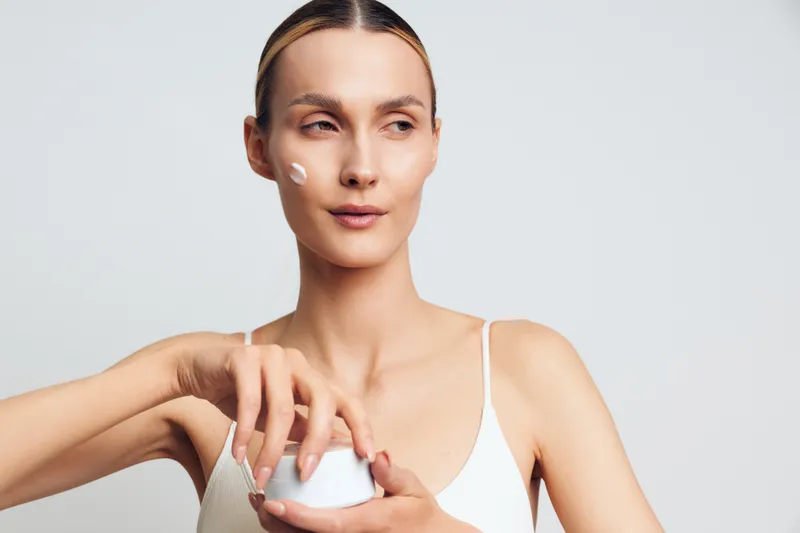 Woman skincare face cream beauty spa portrait, hygiene self care, young woman applies moisturizer on face, calm expression, minimal studio lighting, close up, white background