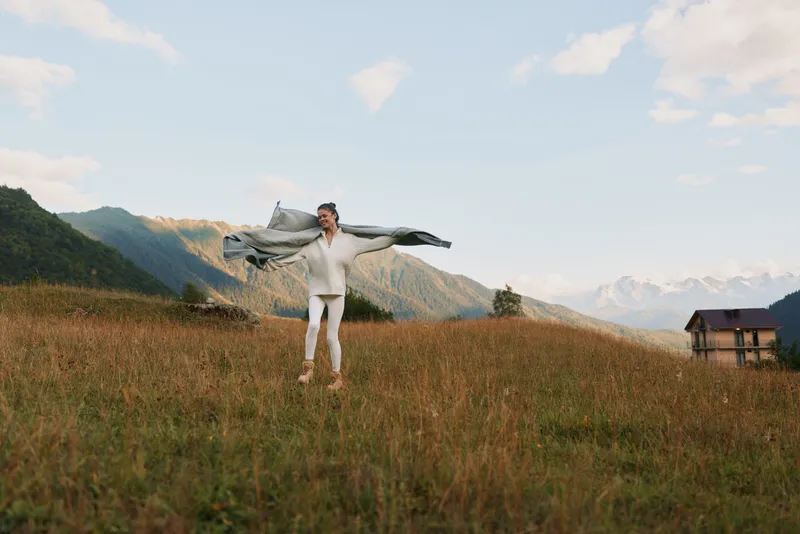 Man in white clothes stands in a grassy field with a scarf spread wide, enjoying the outdoors, bright day, distant mountains, sense of freedom and adventure