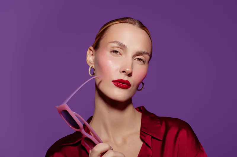 Portrait of confident woman with red lipstick and hoop earrings holding pink sunglasses on purple background. Fashion concept with stylish makeup and elegant red satin blouse.