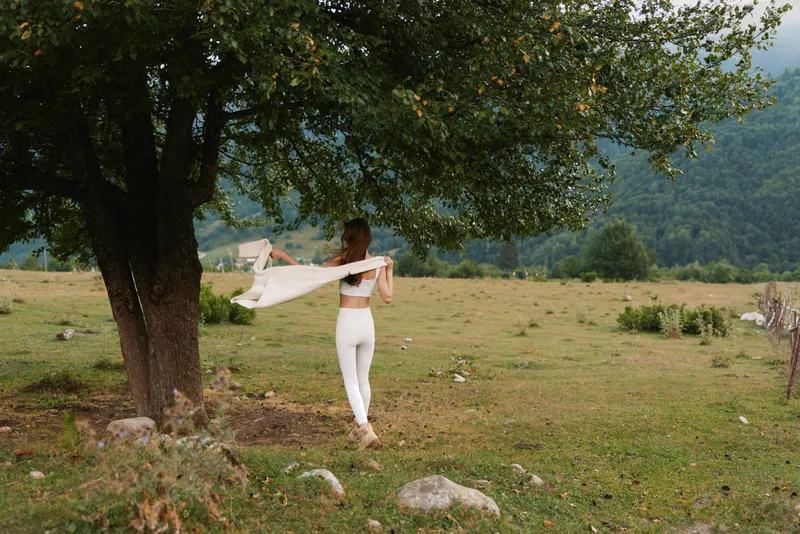 A young woman dances with a flowing scarf beneath a shaded tree in a sunlit countryside field, capturing a carefree, expressive moment of movement and freedom.