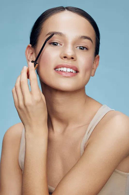Young woman applying eyebrow gel, showcasing natural beauty and confidence against a soft blue background, reflecting beauty, skincare, and self care