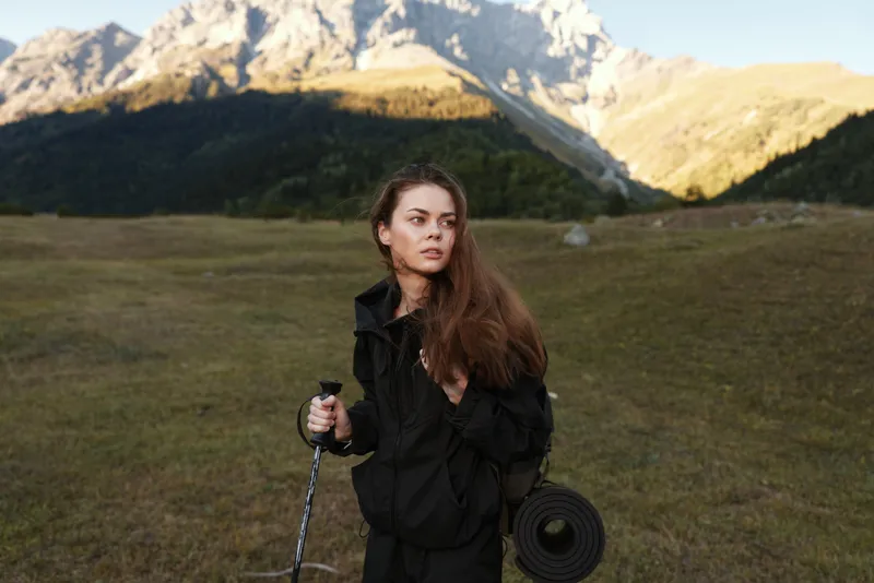 A lone female hiker stands in a vast mountain meadow, adjusting a trekking pole. She wears a dark jacket and carries a rope coil, gazing into the distance with calm determination.