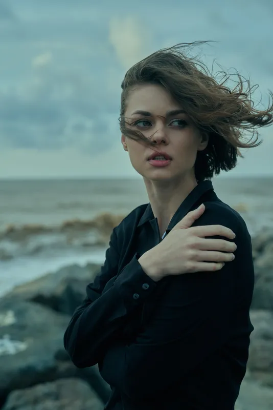 Moody scene of a woman standing by the sea on a windy day, wearing a dark shirt, hair blown across her face, calm waves behind her, capturing resilience and style in natural coastal light