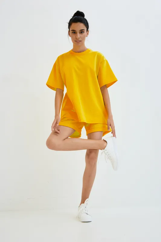 Young woman wearing a yellow oversized t-shirt and shorts poses indoors against a white wall, showcasing casual sportswear with a clean, minimalist background.
