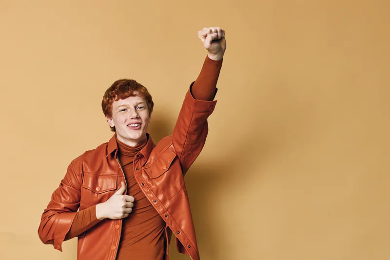 Young man with red hair smiling and raising fist in orange leather jacket and brown turtleneck against beige background with happy confident gesture.