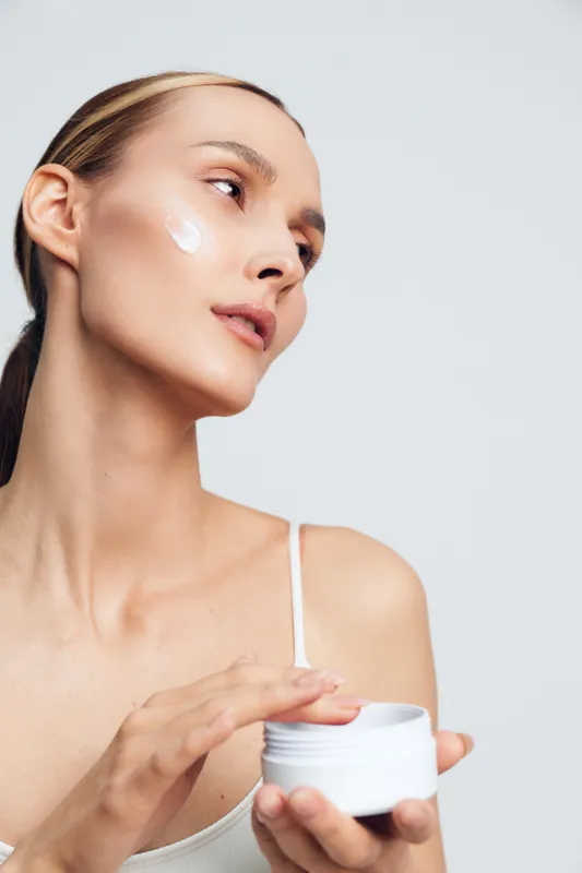 Skincare woman face cream moisturizer beauty jar closeup routine studio portrait She applies white cream from a jar to her cheek, calm look, bright clean background concept personal care