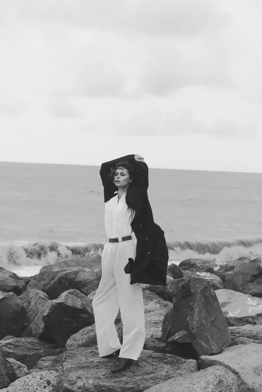 Monochrome scene of a stylish model posing on a rocky seaside, wearing a white jumpsuit and dark coat, arms raised against the wind with a moody horizon for an editorial lifestyle image