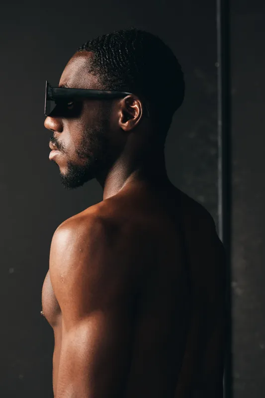 African man with dark skin and muscular body wearing futuristic sunglasses in creative conceptual visual style. Portrait of confident male model with short hair and bare shoulders on dark background.