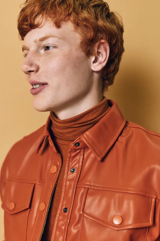 Young man with red hair and fair skin wearing brown leather jacket and turtleneck posing in studio with soft warm lighting and neutral background for fashion portrait.