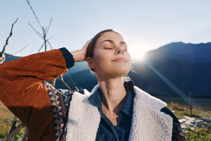 Woman portrait in nature with sunlight and mountains, eyes closed and smiling outdoors, enjoying fresh air and travel vibes in a cozy jacket and serene peaceful mood at golden hour.