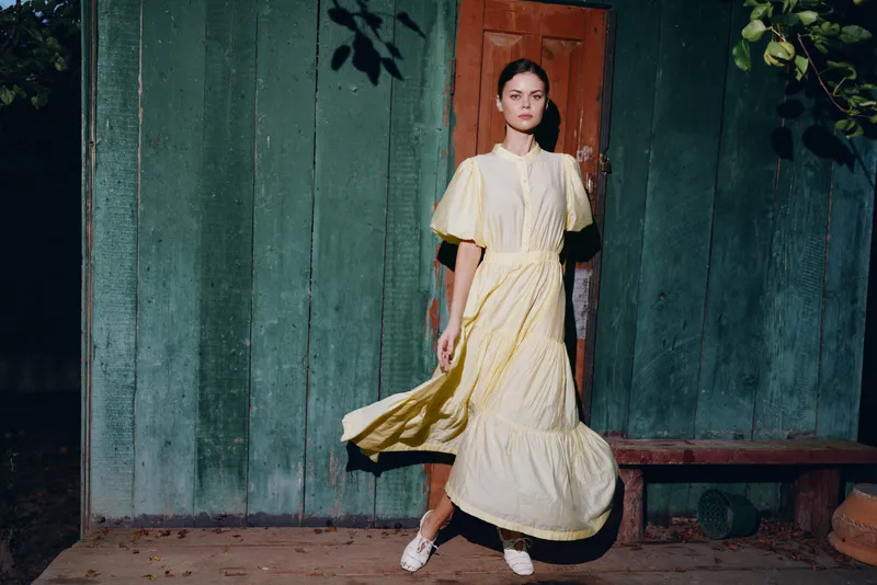 woman wearing a flowing yellow dress walks along a weathered green wall, creating graceful motion in a moody outdoor setting, conveying candid fashion and elegance and confidence