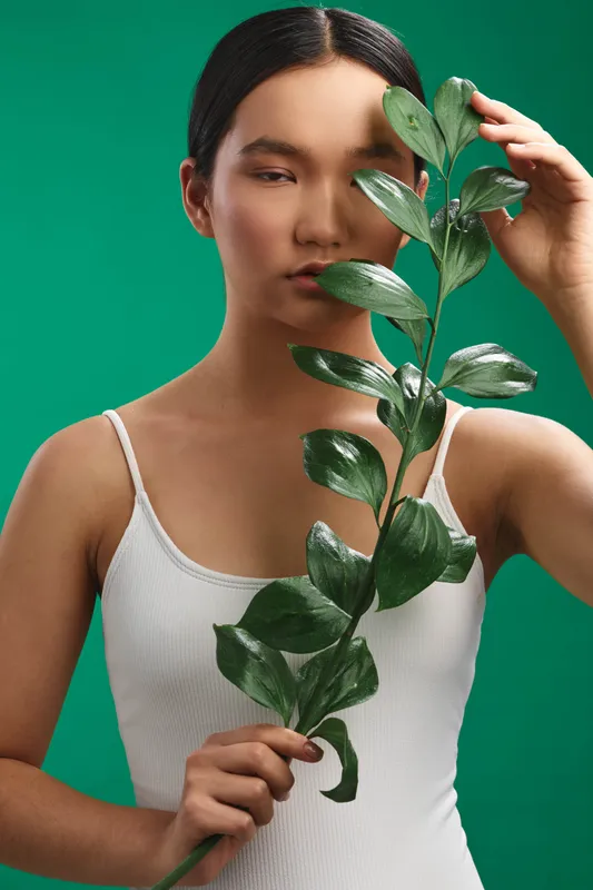 Asian woman holds a leafy stem in a white top, standing before a green studio backdrop. The scene conveys calm, focus, and natural elegance with a minimalist aesthetic.