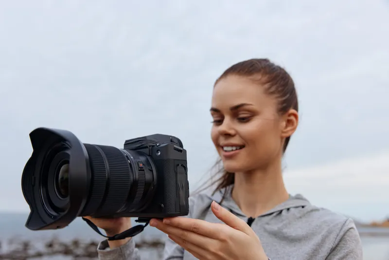 Young woman joyfully holding a camera on a beach, dressed in casual gray attire, with a serene ocean backdrop, embodying creativity and passion for photography