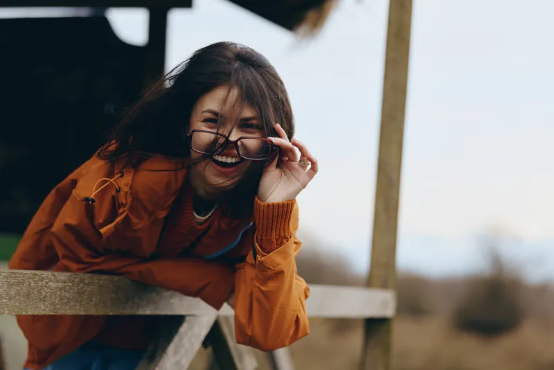 Woman lifestyle portrait outdoors in winter, wearing orange coat and glasses, leaning on wooden railing, smiling while adjusting spectacles, soft daylight, blurred background, candid
