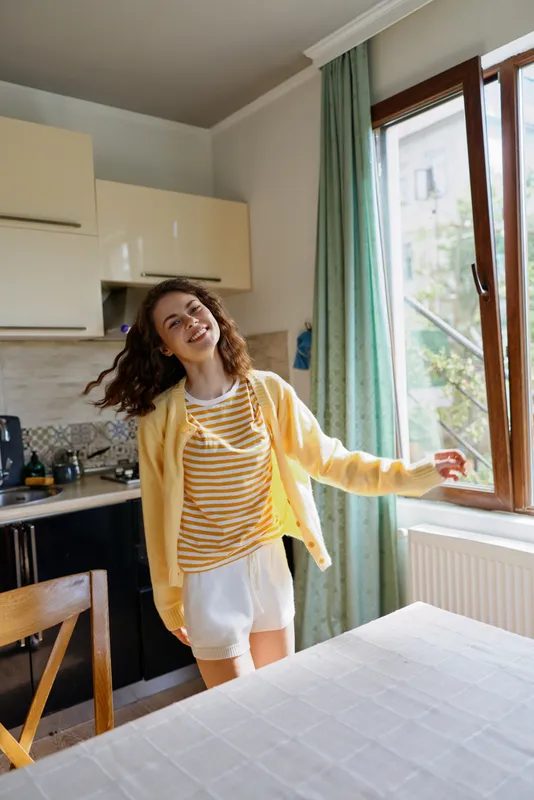 Cheerful woman indoors in a cozy kitchen, wearing a striped shirt and yellow cardigan, enjoying daylight through a large window, conveying relaxed home lifestyle and sunny mood