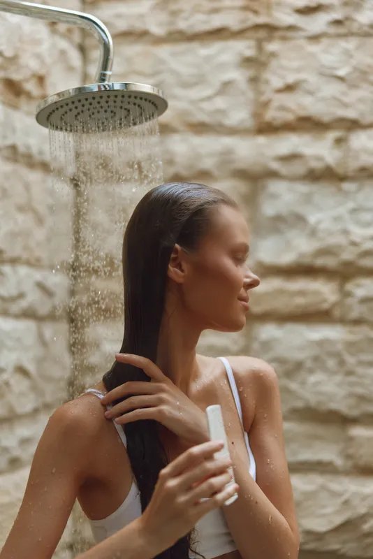 Woman taking an outdoor shower with wet hair against a textured stone wall, enjoying a refreshing moment of relaxation and natural skincare routine.