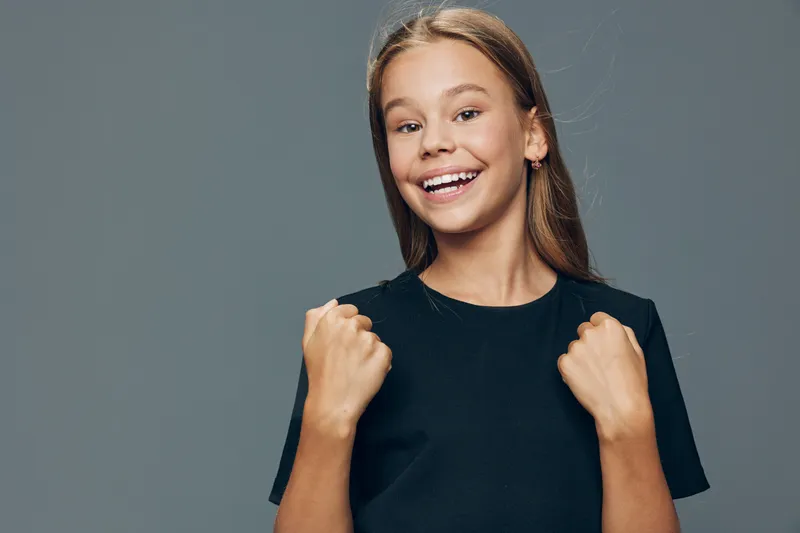 Teenage girl with long hair smiling and showing fists for success or celebration on gray background. Youth, happiness, empowerment concept with casual style and cheerful emotion.