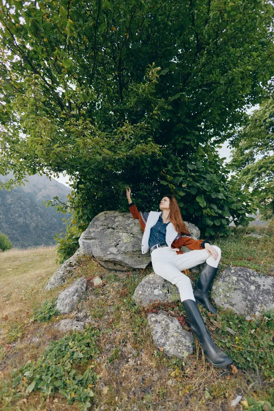 Woman in boots sitting on rocks under a tree in a meadow, relax in countryside nature. Casual fashion jacket and scarf portrait on hillside with green foliage and peaceful landscape.