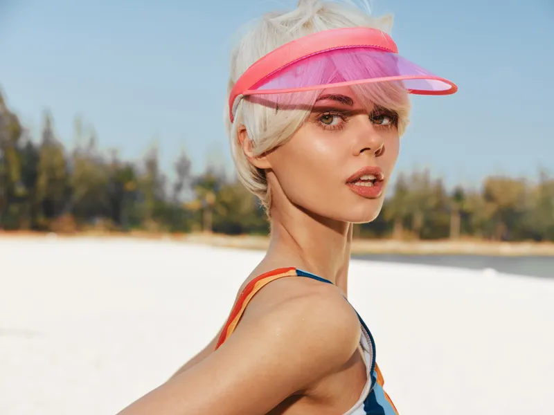 A stylish woman on a bright beach wears a pink visor, sunlit skin, and playful summer attire, turning toward the camera with a confident, fashionable gaze.