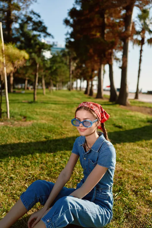 Lifestyle woman in modern boho-western denim street style sitting on grass in sunny park wearing blue sunglasses and red bandana with film color tones enhancing the mood.