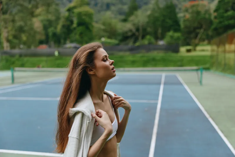 Young athletic woman in sportswear enjoying a moment of relaxation on a tennis court, showcasing her fitness and confidence against a lush green background