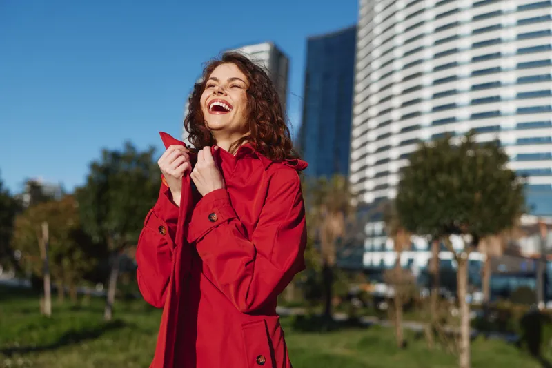 A young woman in a red coat laughs in a sunny city park, with tall modern buildings rising behind her, conveying carefree urban happiness and optimism.