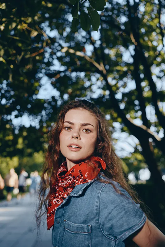 Lifestyle woman in modern boho-western denim street style with a red bandana, film color tone, natural light, and relaxed outdoor urban setting with green foliage.