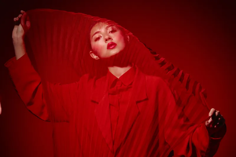 Young woman in red outerwear posing with sheer red fabric. Bright lip color and makeup highlight her face. Fashion and beauty shot with intense red lighting for creative concept.