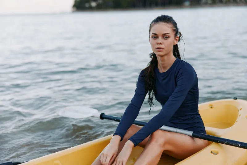 Female rower sits in a yellow canoe on calm water, wearing a navy long sleeve top. She gazes ahead with composed expression, ready to paddle, embodying focus and outdoor adventure.