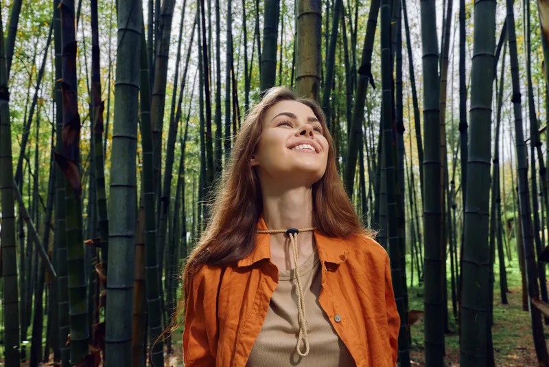 Woman in bamboo forest smiles and looks upward, enjoying nature outdoors in a green grove. Young female portrait wearing orange jacket, peaceful, serene and relaxed mood in woods.