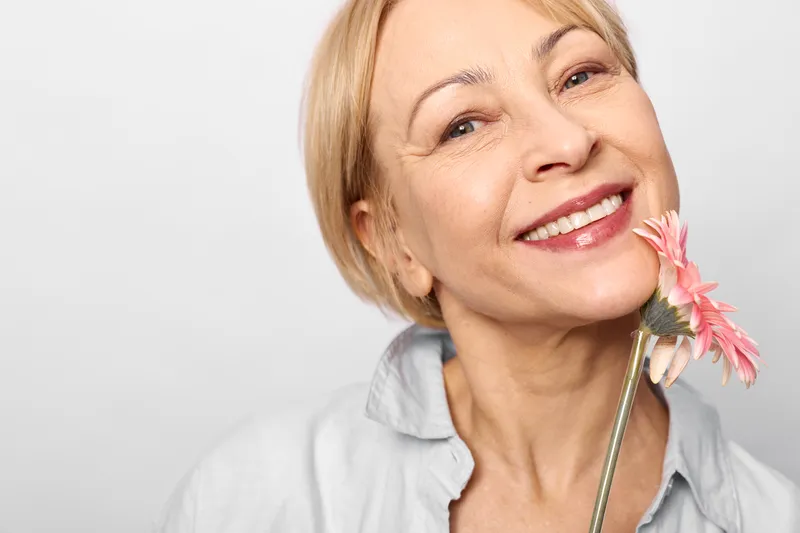 woman smiling older closeup flower beauty skincare wellness portrait studio, smiling older woman holds a pink flower by her cheek, close up in a white studio background, beauty care wellness