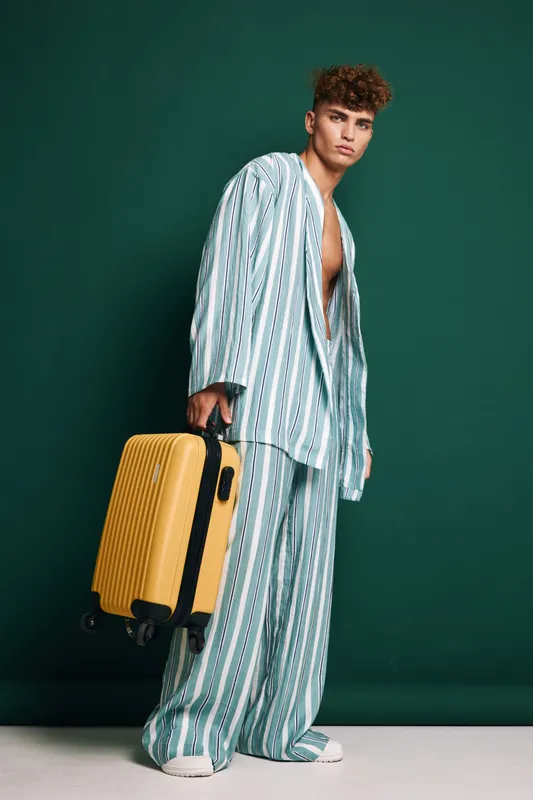 Young man in striped pajamas holding yellow suitcase standing against green background for travel, fashion, and lifestyle concept with casual outfit and luggage.