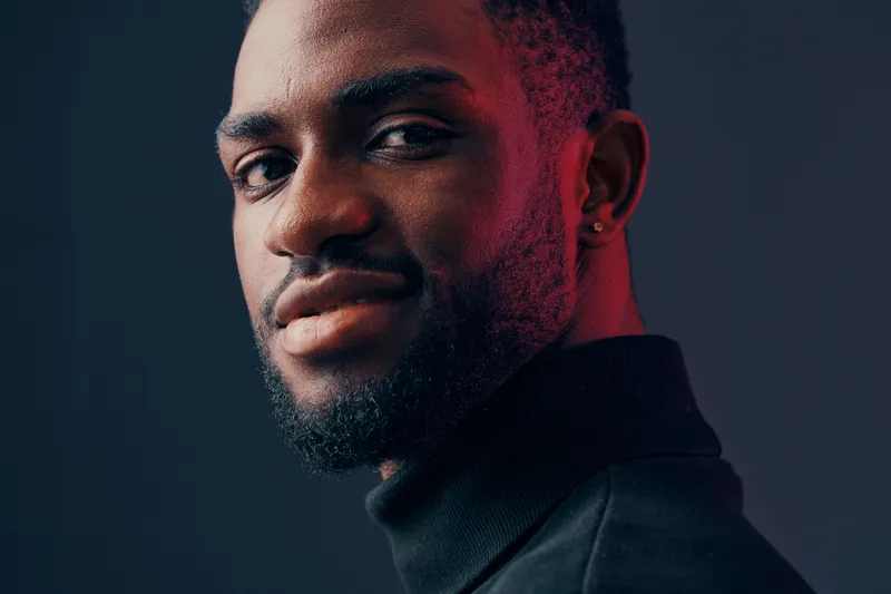 Stylish African male model in a turtleneck sweater, showcasing confidence and charisma against a dark, moody backdrop with vibrant lighting