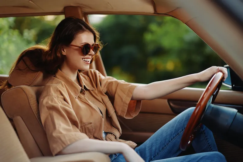 Young woman driving a vintage car, smiling joyfully, wearing sunglasses and a casual outfit, with a blurred green background, reflecting a carefree summer vibe.