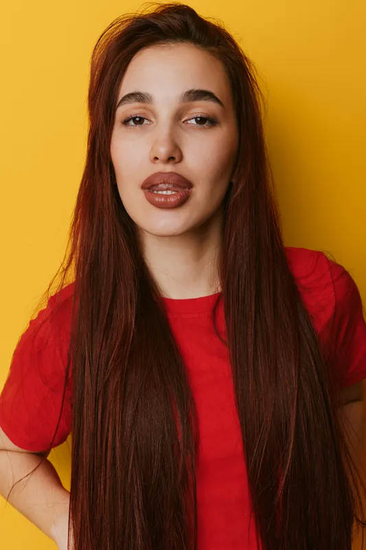 Young woman with long straight hair and full lips wearing red shirt posing against yellow background with confident expression and hand on hip in studio portrait.
