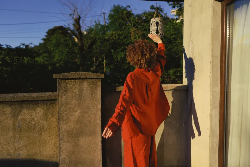 Curly haired woman in an orange dress reaches toward a hanging lantern on a sunlit balcony, capturing a candid moment of curiosity and calm amid warm tones and quiet afternoon light
