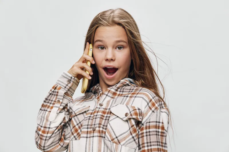 Girl talking on phone with surprised expression wearing plaid shirt on white background. Child communication, mobile device, casual clothing, emotions and lifestyle portrait indoors.