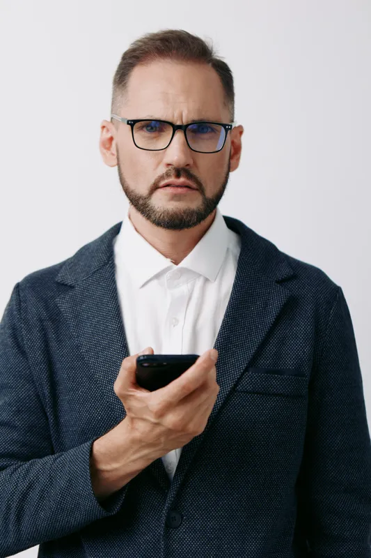 A confident business professional man on an isolated colored background, wearing blazer and glasses, with a serious expression, holding a smartphone and ready for strategic communication.