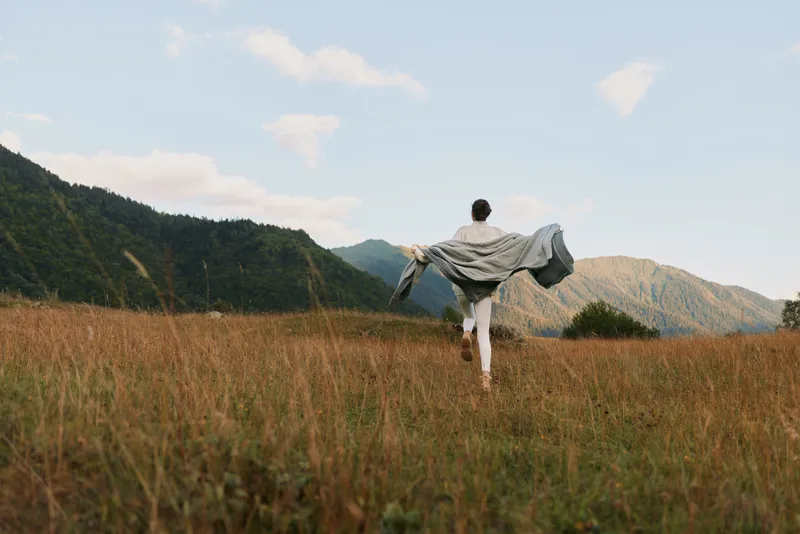 Young woman in white pants walks through a dry grass field holding a blanket behind her with mountains in the background under a partly cloudy sky.