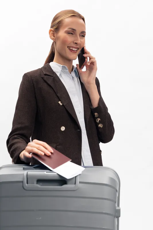 Smiling woman in business attire holds passport and boarding pass while talking on phone near suitcase. Concept for travel, business trip, communication and modern lifestyle.