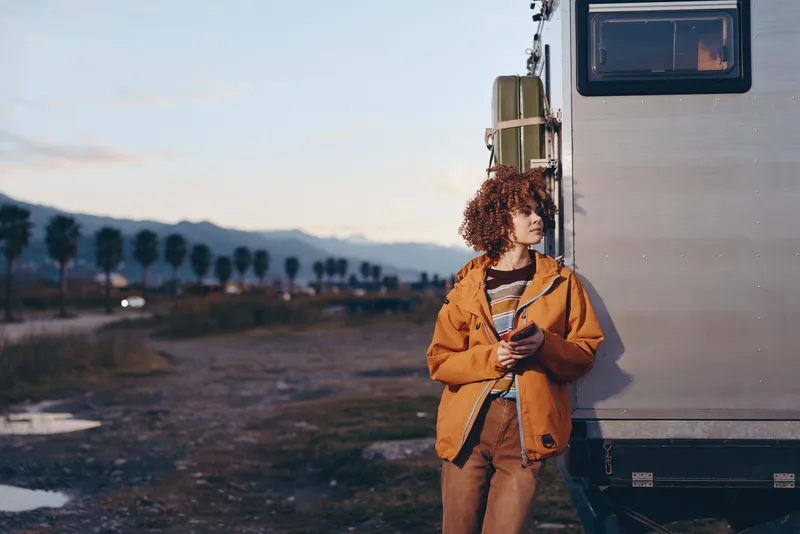 Woman lifestyle outdoors beside a travel van at sunset, curly haired woman in orange jacket holding a backpack strap, warm light over mountains, calm adventure mood, road trip, sunrise