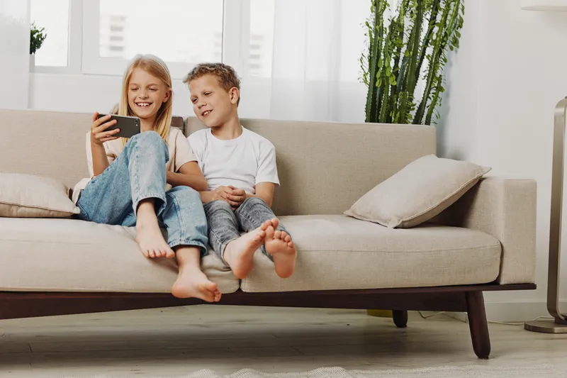 Two happy children sitting barefoot on beige sofa watching smartphone, smiling and relaxing in bright living room with modern decor, casual clothes and natural light.